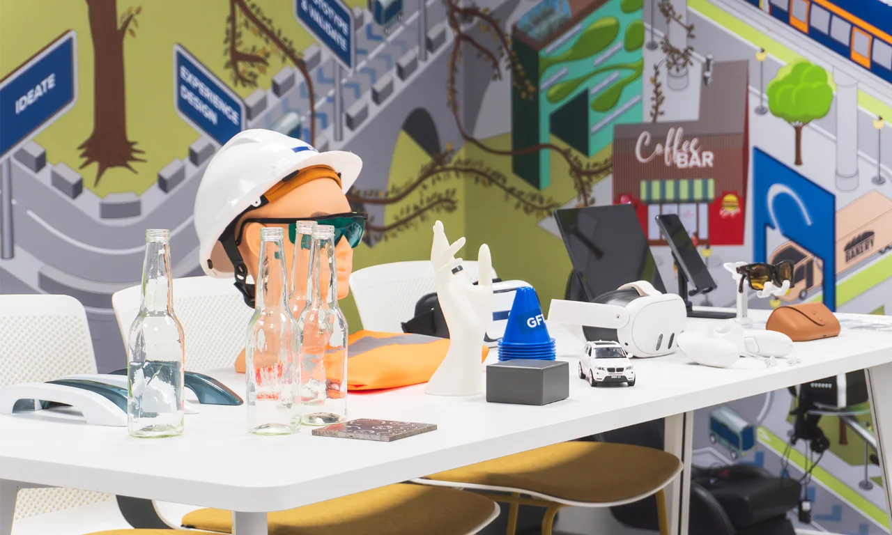 This image showcases a table in an innovation lab, adorned with an array of technological gadgets and equipment. Items on the table include a VR headset, a robotic hand model, a hard hat with safety glasses, and several empty glass bottles. The backdrop features a vibrant mural depicting various innovation-related themes, adding to the creative and futuristic ambiance. The setting emphasizes the lab&#039;s focus on exploring and developing new technologies and solutions.
