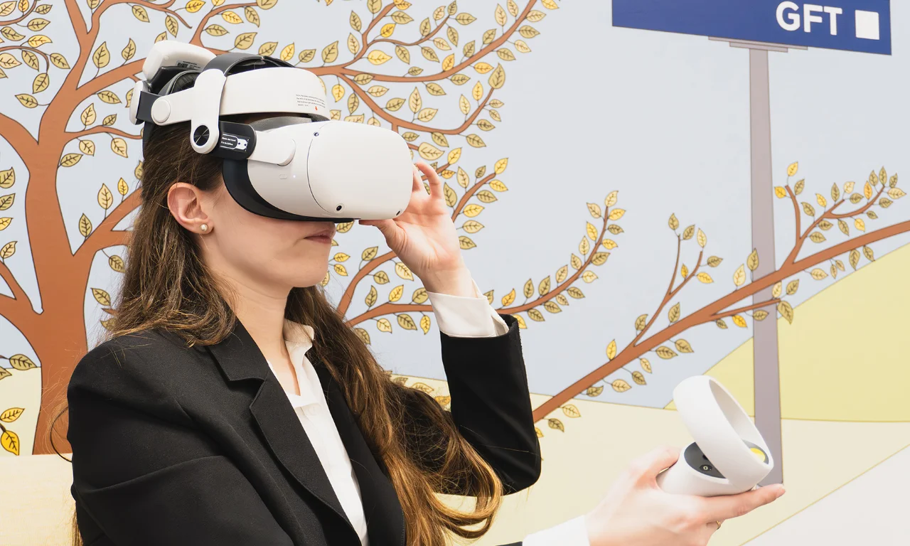 This image depicts a woman immersed in a virtual reality (VR) experience within an innovation lab. She is wearing a modern VR headset and holding a VR controller, fully engaged in the virtual environment. The background features a creative and colorful mural with a tree and the GFT logo, highlighting the innovative and tech-focused atmosphere of the lab. The setting emphasizes the use of advanced VR technology for immersive learning, training, or simulation purposes.
