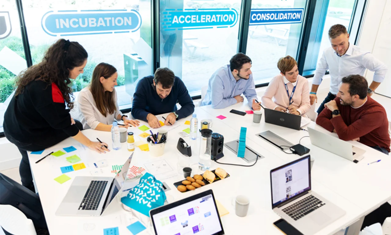 This image depicts a dynamic co-creation session in an innovation lab. A diverse group of professionals is actively engaged in a brainstorming session around a large table. The workspace is equipped with laptops, sticky notes, markers, and other office supplies, creating an environment conducive to creativity and collaboration. The glass windows in the background feature motivational words like &quot;Incubation,&quot; &quot;Acceleration,&quot; and &quot;Consolidation,&quot; emphasizing the stages of innovative development. The team members are deep in discussion, jotting down ideas, and working together to solve problems, demonstrating the essence of collaborative innovation.