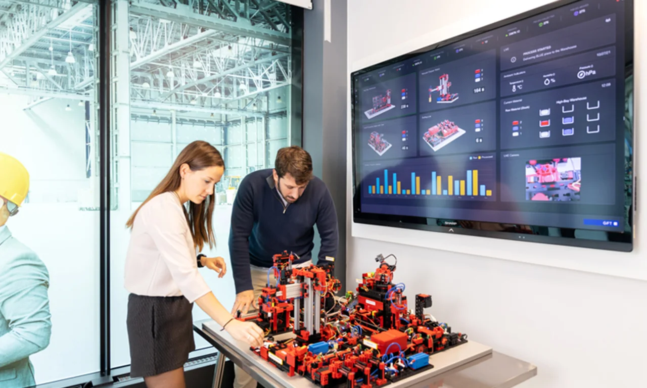 This image showcases a collaborative session in an innovation lab focused on industrial automation. A woman and a man are engaged with a sophisticated setup of automation equipment, featuring a detailed, miniaturized industrial model with numerous components and wiring. A large screen on the wall displays real-time data analytics and visualizations related to their project, illustrating various metrics and system statuses. The backdrop includes a spacious, modern industrial setting visible through large windows, emphasizing the lab&#039;s integration with real-world industrial applications. This environment highlights the synergy between hands-on experimentation and advanced data-driven insights, fostering innovation in industrial technology.