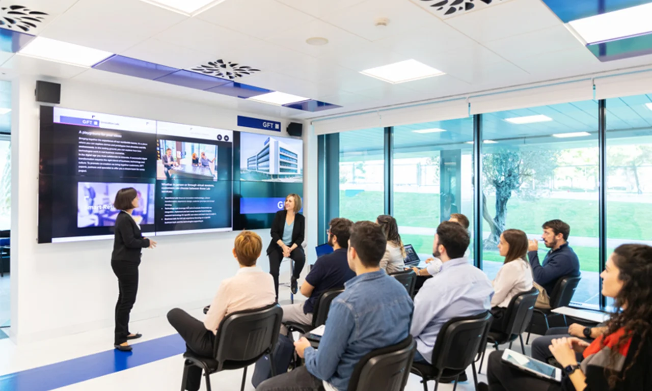 This image captures an interactive presentation taking place in an innovation lab auditorium. The presenter is standing in front of a large digital screen, displaying various multimedia content to an attentive audience. The attendees are seated in rows, equipped with laptops and tablets, ready to take notes and participate. The room is bright and modern, with large windows providing a view of a green outdoor area, enhancing the open and collaborative atmosphere. This setup fosters engagement and knowledge sharing, essential for innovative thinking and development.