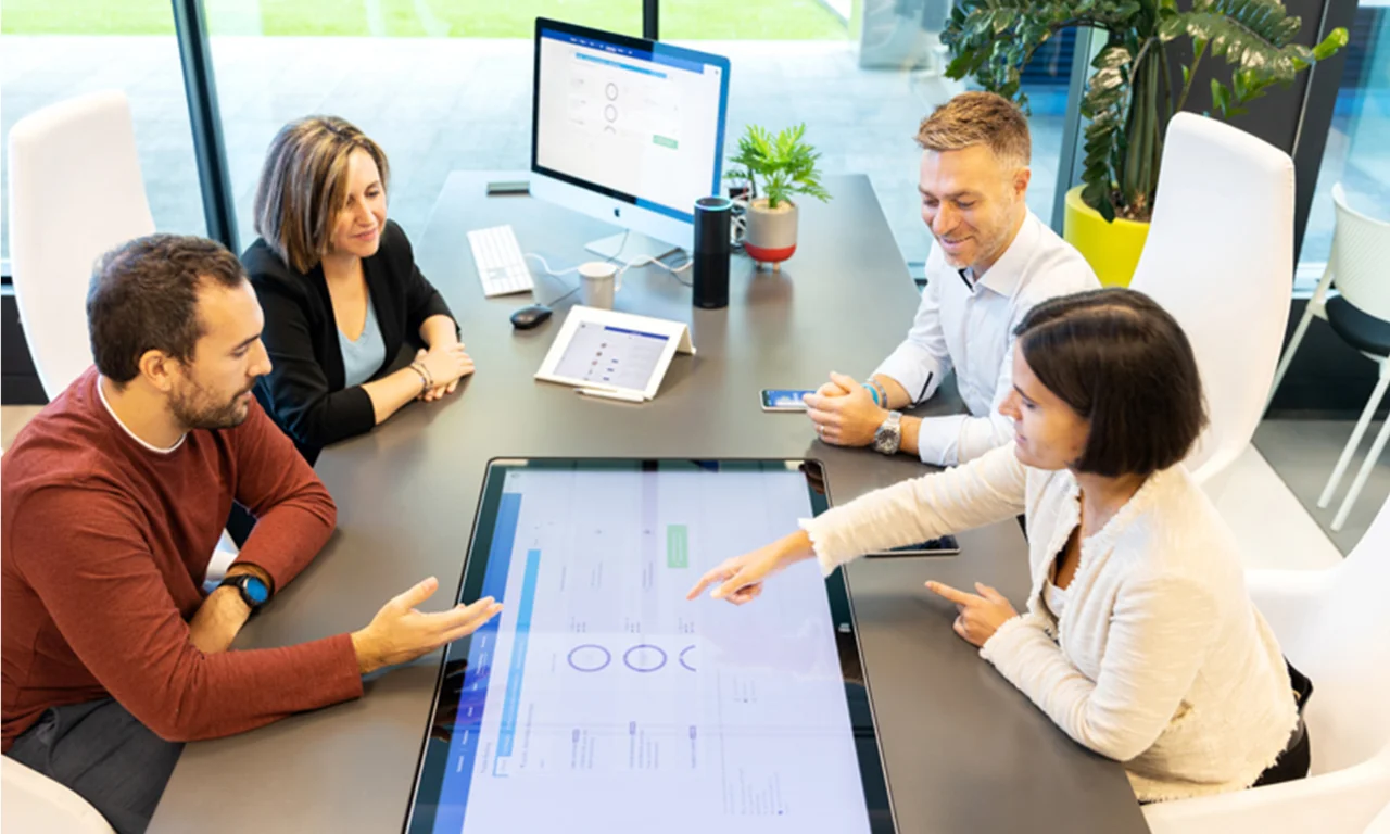 This image shows a collaborative meeting in an innovation lab at a bank branch. Four team members are seated around a modern table, engaging with a large interactive touchscreen display embedded in the table. They are discussing data and insights displayed on the screen. Additional digital devices, such as a tablet, smartphone, and desktop computer, are also in use, highlighting the integration of technology in their workflow. The bright, open space with large windows and indoor plants creates an environment conducive to innovative thinking and teamwork.
