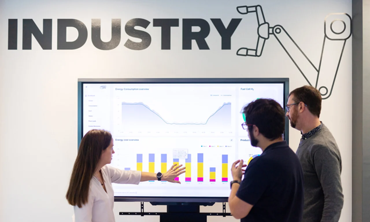 Esta imagen muestra un espacio de laboratorio de innovación con temática industrial en el que tres profesionales participan en una discusión. Están concentrados en una gran pantalla digital que muestra varios gráficos y visualizaciones de datos relacionados con el consumo y los costos de energía. La pantalla es parte de una configuración moderna diseñada para facilitar el análisis y la colaboración. El entorno se ve realzado por un destacado cartel con la palabra &quot;INDUSTRIA&quot; y un gráfico de un brazo robótico en la pared, lo que enfatiza el enfoque industrial y tecnológico del laboratorio. Este entorno resalta la dedicación del laboratorio de innovación a la integración de tecnología avanzada y conocimientos basados en datos en procesos y soluciones industriales.