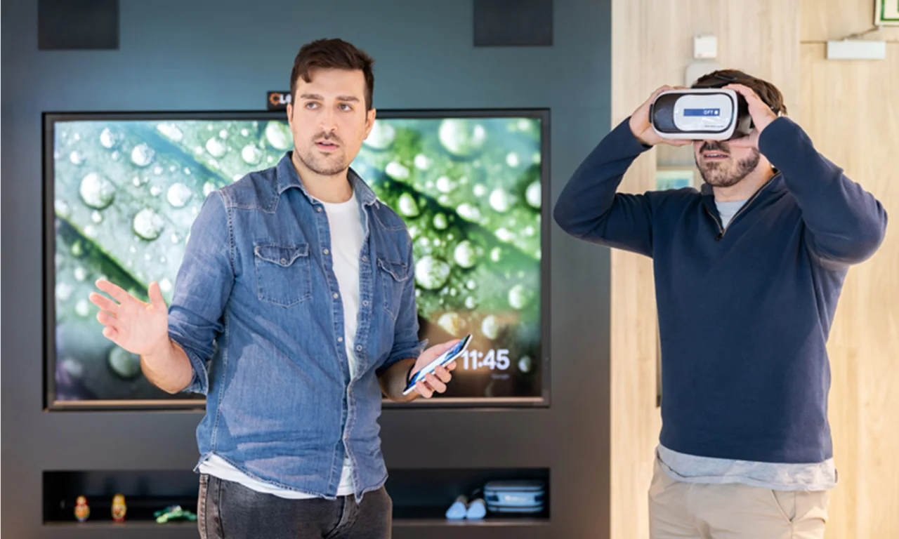 Esta imagem captura uma sessão interativa em um laboratório de inovação onde dois homens estão interagindo com tecnologias avançadas. Um homem está usando um headset de realidade virtual, experimentando conteúdo imersivo, enquanto o outro homem está segurando um smartphone e possivelmente explicando ou apresentando algo. O fundo apresenta uma tela grande exibindo visuais vívidos, aprimorando o ambiente moderno e centrado em tecnologia do laboratório de inovação. Este cenário reflete a integração de tecnologias de ponta e ambientes colaborativos voltados para promover a inovação e a criatividade.