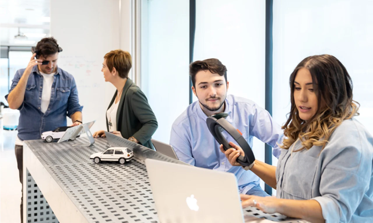 Esta imagem captura uma cena dinâmica em um laboratório de inovação onde uma equipe de profissionais está envolvida em atividades colaborativas. Um homem à esquerda está usando óculos de realidade aumentada (RA), indicando um ambiente de alta tecnologia. Uma mulher e outro homem estão interagindo com modelos em escala de veículos em uma mesa de metal perfurada, sugerindo um foco na tecnologia automotiva. Em primeiro plano, uma mulher está usando um laptop enquanto segura um scanner portátil, destacando a integração de ferramentas digitais e protótipos físicos. O ambiente de laboratório moderno e brilhante com grandes janelas e quadros brancos ao fundo promove a criatividade e a inovação na indústria automotiva.